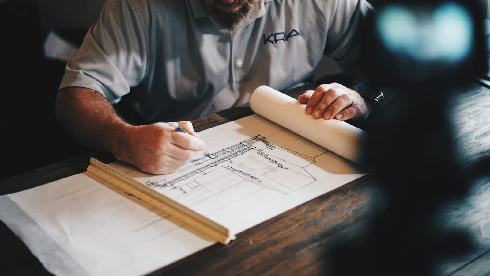 A man wearing a 'KRA' shirt drafts detailed architectural plans with a pen.